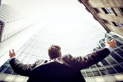 Businessman with arms raised looking up at skyscrapers.