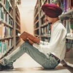 Man in turban sitting on library floor reading a book between bookshelves.
