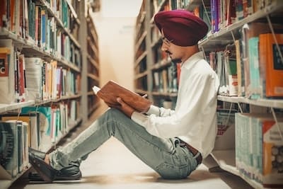 Man in turban sitting on library floor reading a book between bookshelves.