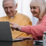 Two elderly individuals smiling while looking at a laptop screen together.